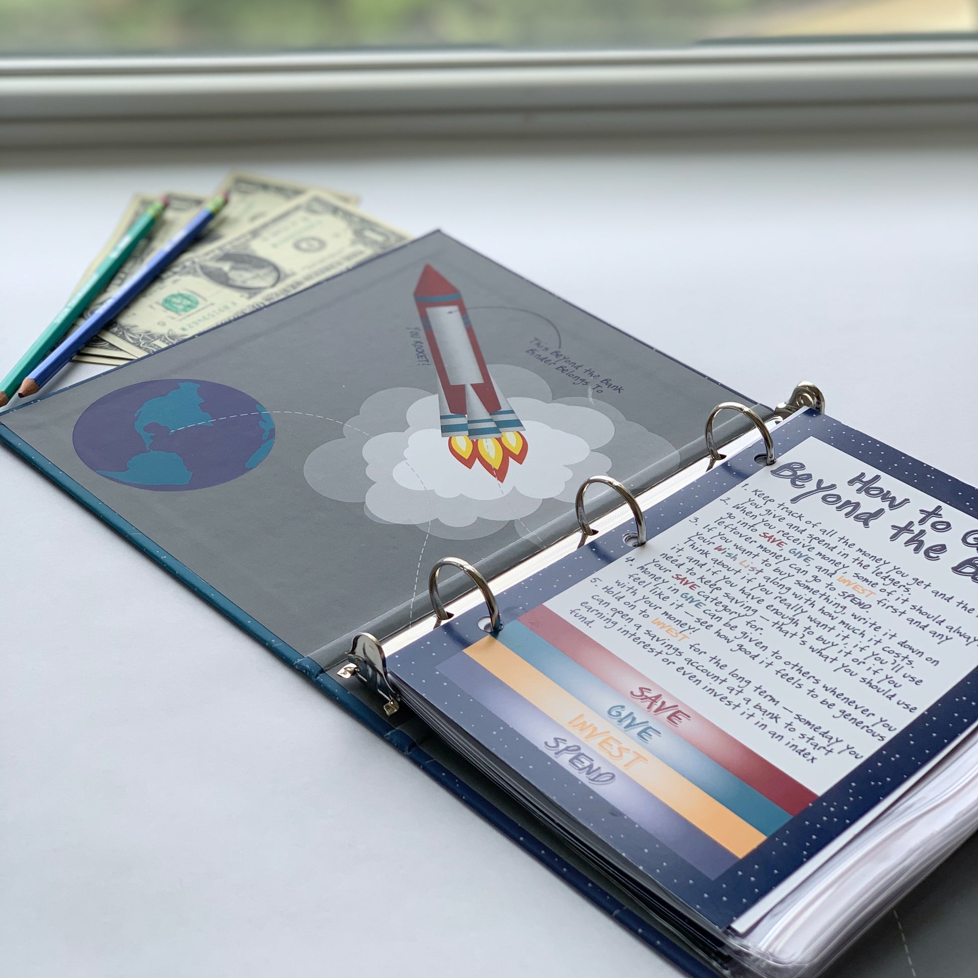 image of space themed money management binder open to front page with dollar bills and pencils next to it